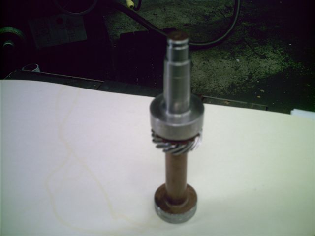 Metal shaft with a helical gear and two circular flanges, resting on a white surface