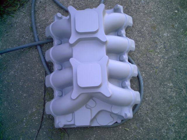 Primer-gray dual-quad intake manifold with two carb pads sitting on concrete