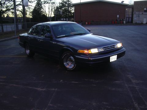 1996 Ford Crown Victoria - A dark blue sedan is parked in an asphalt lot with a building and trees in the background.