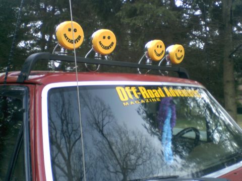 1989 Ford Bronco - A red SUV is equipped with a roof rack holding four yellow lights smiley faces and an "Off-Road Adventure" magazine sticker.