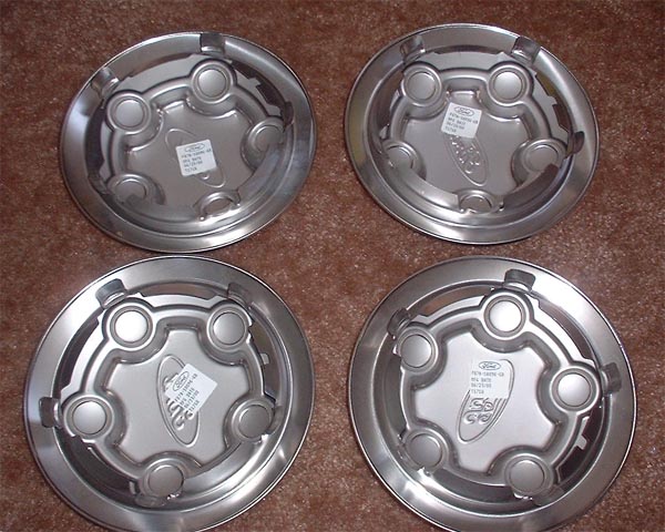 Four Ford hubcaps on a carpeted surface. One has a sticker with Ford branding and part numbers