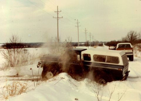 1979 Ford Bronco - A pickup truck with a camper shell is stuck in the snow on a rural road, with another truck visible in the background.