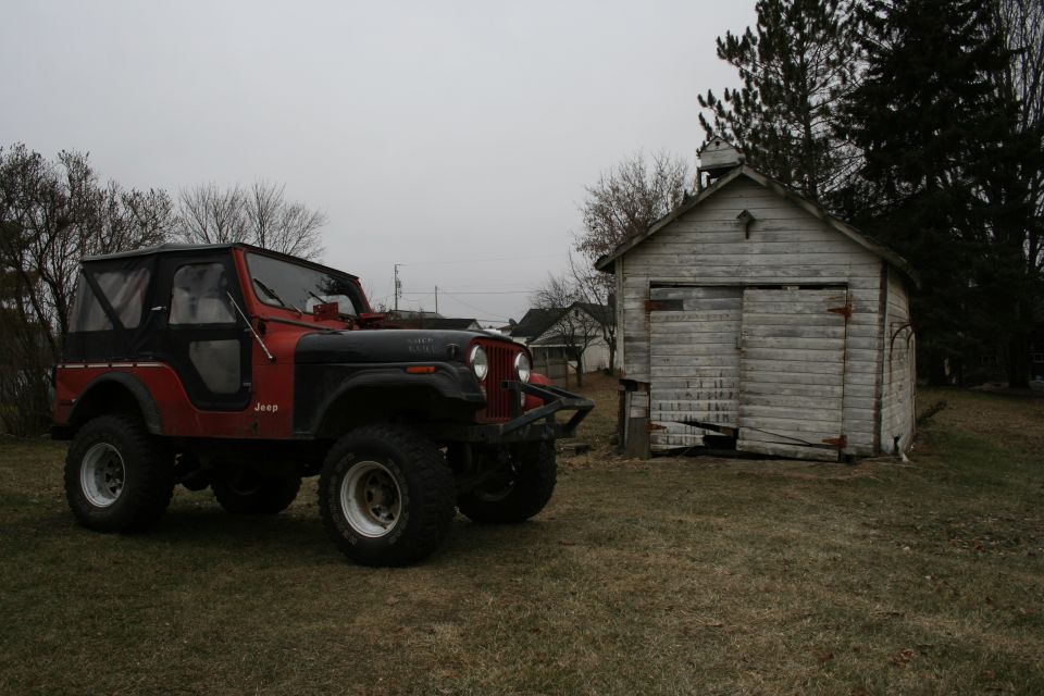 Red Jeep CJ with black soft top on lifted mud tires parked beside a weathered shed