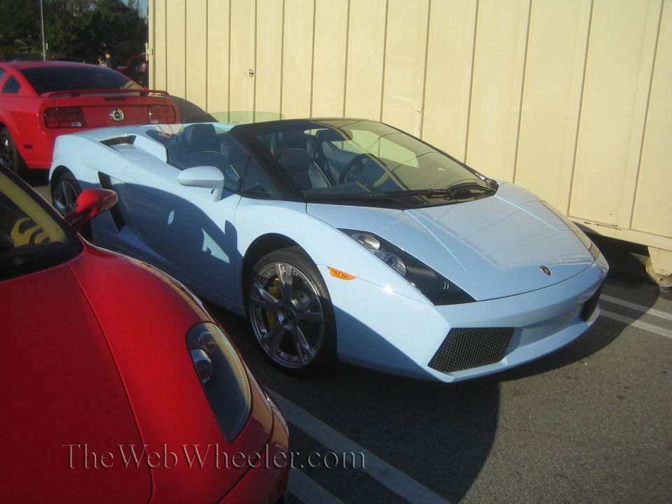 Light blue convertible sports car parked next to a red car, with a red Mustang in the background