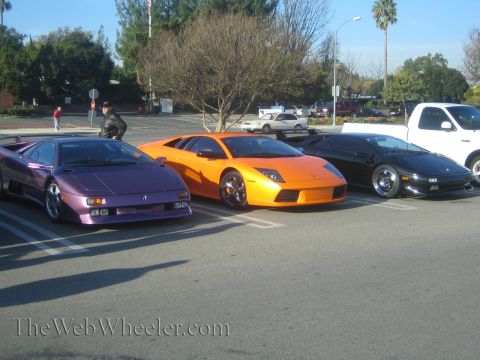 1985 Ford Mustang - Three exotic sports cars are parked side-by-side in a parking lot on a sunny day.