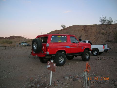 1985 Ford Bronco - A red Ford Bronco is parked in a desert landscape next to a white pickup truck, with a sign reading '20 E 19' in the foreground.