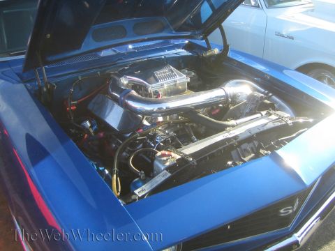 1985 Ford Mustang - Engine bay of a blue muscle car at a car show, with another vehicle in the background.
