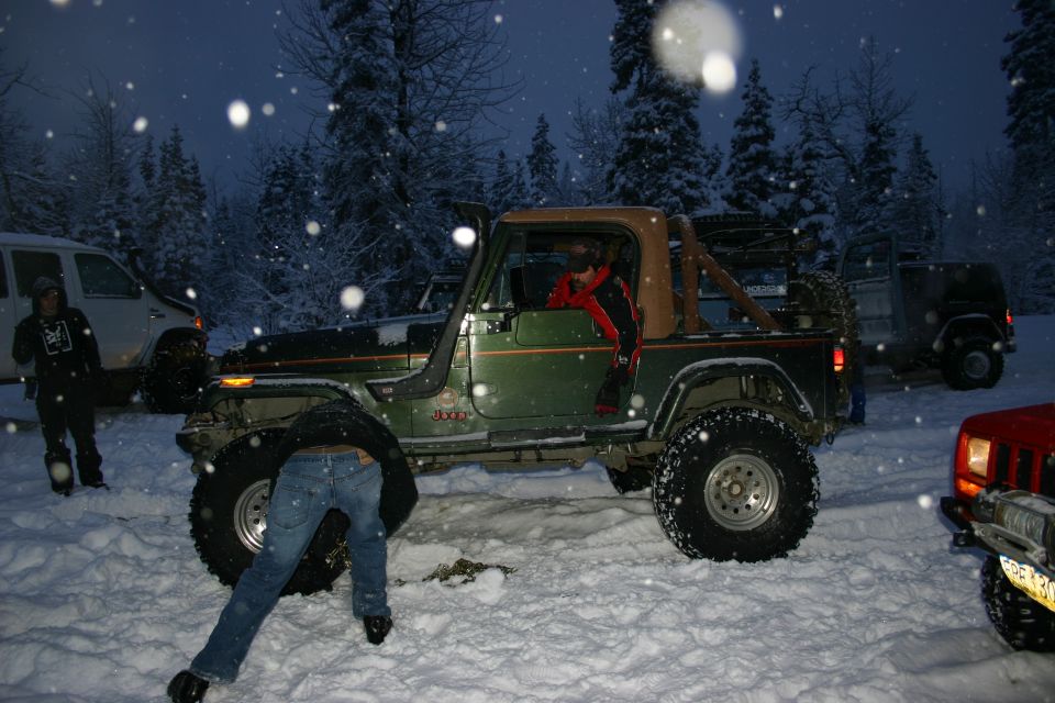 Green Jeep Wrangler YJ with a person underneath, in the snow. Other vehicles visible