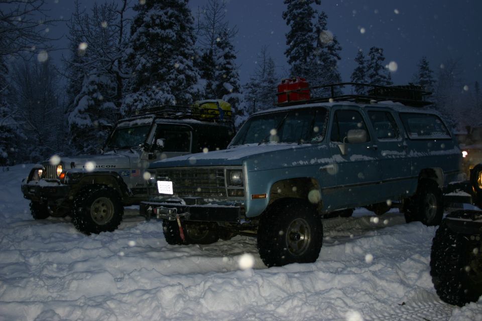 Jeep Wrangler and blue Chevrolet Suburban parked in snow, trees in background