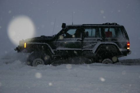 2003 Jeep TJ - A dark green Jeep Cherokee XJ navigates through deep snow, with visible text on its side indicating "AlaskaSkyParts.com".