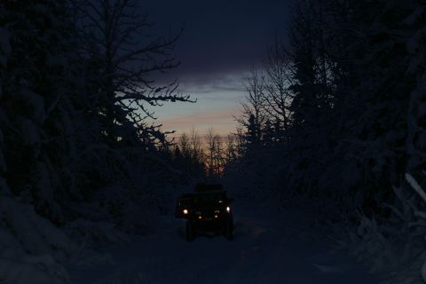 2003 Jeep TJ - A dark SUV drives down a snow-covered road surrounded by trees as the sun sets, creating a dramatic sky.