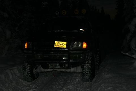 2003 Jeep TJ - A black Jeep TJ with a yellow license plate drives on a snowy path at night, illuminated by its headlights and auxiliary lights.