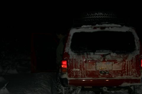 2003 Jeep TJ - A red Jeep SUV is parked in the snow at night, with a spare tire mounted on the roof rack.