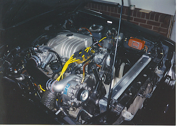 Engine bay of a black 1986 Ford Mustang GT with a supercharger and yellow spark plug wires