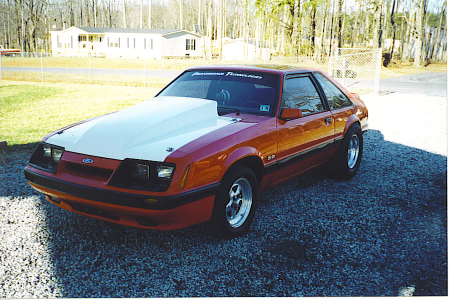 Orange Ford Mustang with a white cowl induction hood and "5.0" badge on the fender