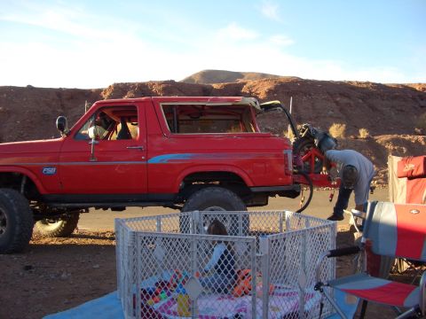 1985 Ford Bronco - A red 1980s Ford Bronco is parked outdoors with a person working on the rear tire carrier.