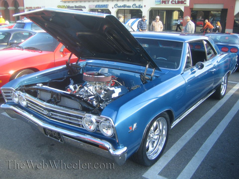 Blue classic car, hood up, showing engine. Another red car is partially visible to the left