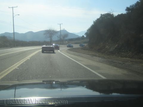 1985 Ford Mustang - Several cars drive along a highway with hills and mountains visible in the distance.