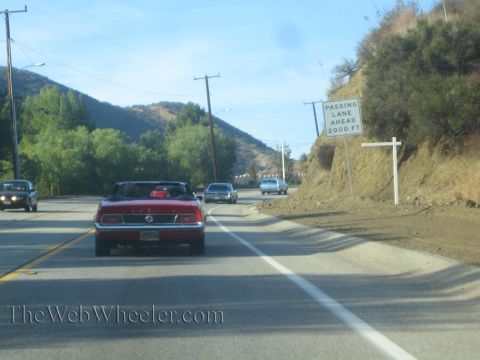 1985 Ford Mustang - A red convertible, likely a 1969 model, drives down a highway with other vehicles.
