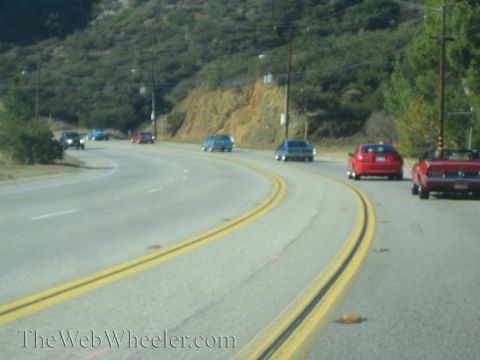 1985 Ford Mustang - A group of cars travel along a scenic route on a sunny day.