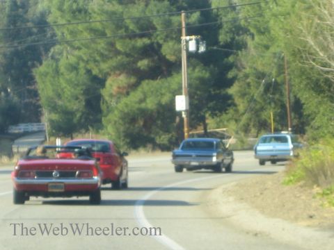 1985 Ford Mustang - A red convertible is seen driving on a road with other vehicles.