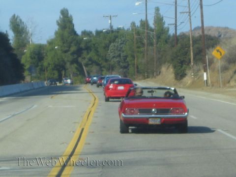 1985 Ford Mustang - A line of cars travels down a two-lane road, led by a red convertible.