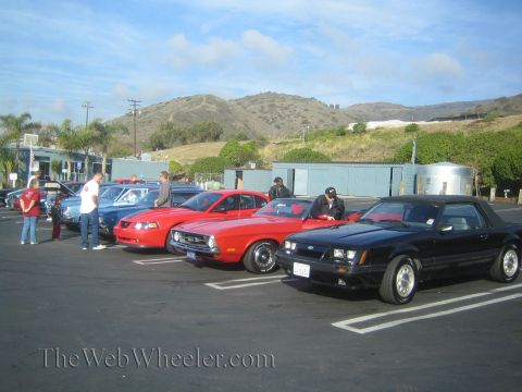 1985 Ford Mustang - Cars parked in a lot with people milling around, under a partly cloudy sky.