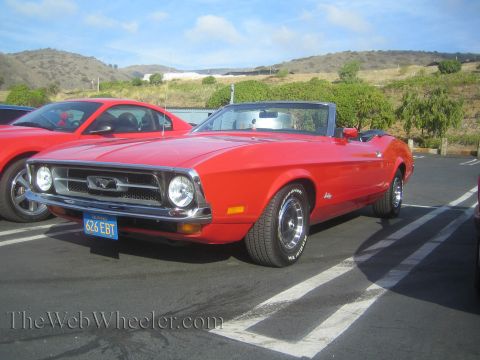 1985 Ford Mustang - This red convertible, likely from the early 1970s, is parked next to another red vehicle at a car show.