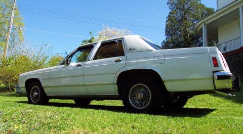1985 Ford Crown Victoria LTD - This white sedan is parked on a grassy lawn with a house in the background under a clear blue sky.