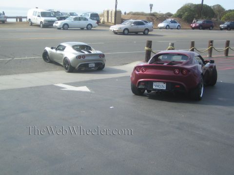 1985 Ford Mustang - Two Lotus Elise sports cars are parked near a coastal road, with other vehicles visible in the background.