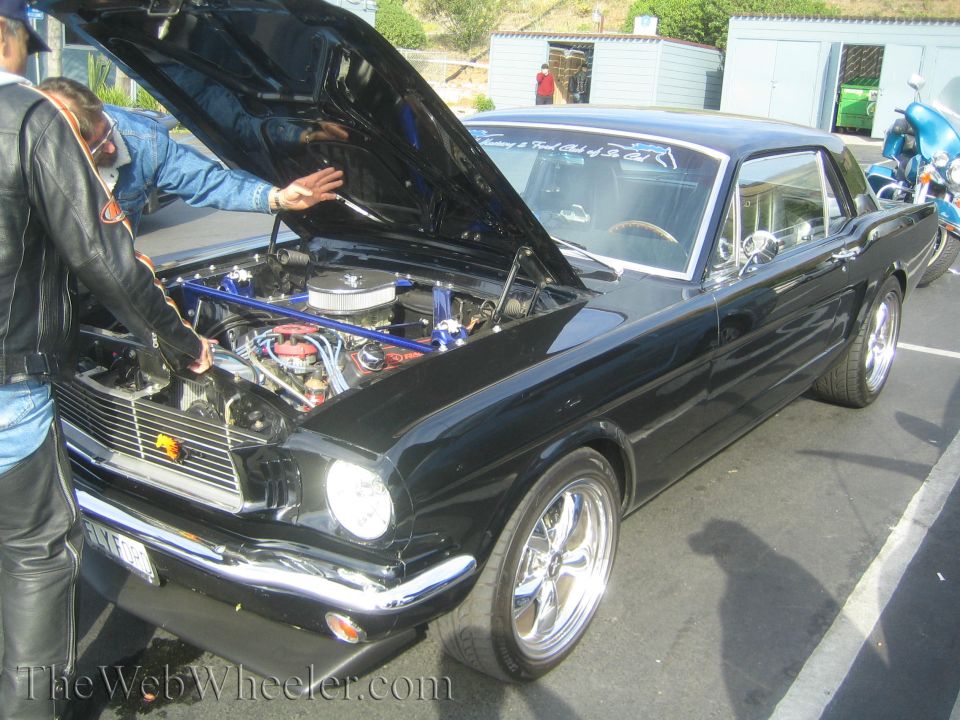 Black coupe with hood open, showing engine and chrome wheels. Two people are looking at the engine