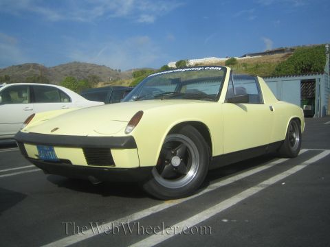 1985 Ford Mustang - A yellow Porsche 914 is parked in a lot, with a white car to its left and a blue sky above.