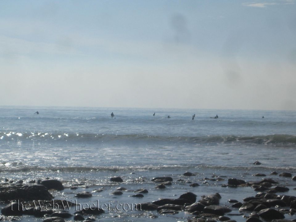 Ocean scene with surfers and rocks in the foreground, watermarked with 'TheWebWheeler.com'