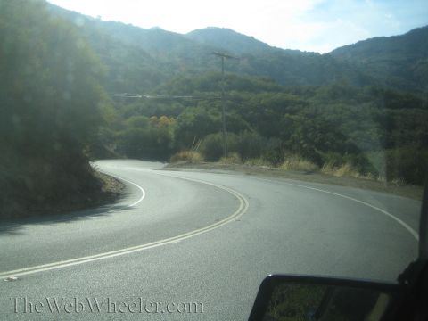 1985 Ford Mustang - A scenic view of a winding road through a mountainous, tree-covered landscape, with a car's side mirror visible.