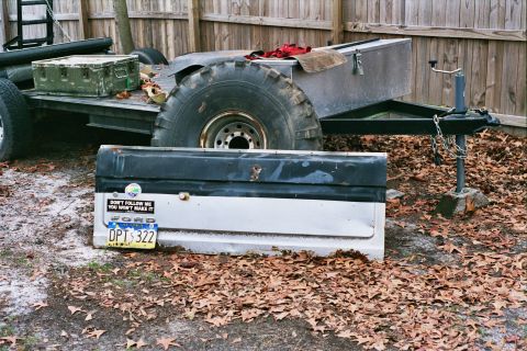 1978 Ford Bronco - A detached Ford tailgate rests on fallen leaves near a utility trailer.