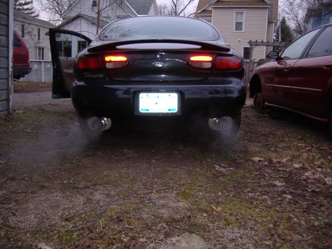 1998 Ford Taurus - A black sedan emits smoke from its dual exhaust pipes, with a red car visible to the side.