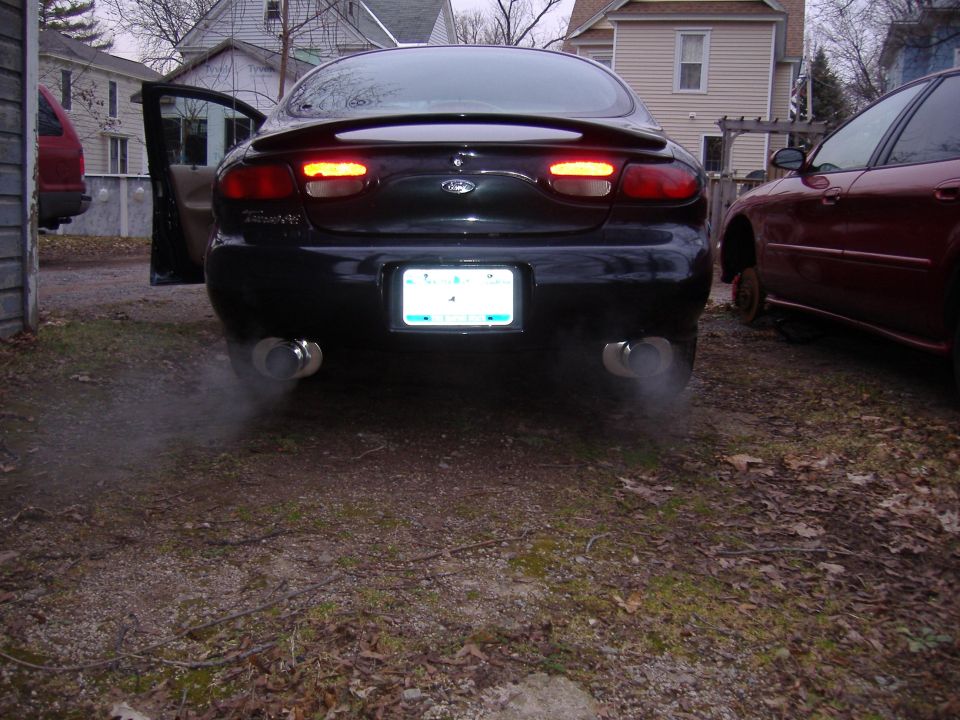 black sedan with exhaust smoke coming from dual exhaust pipes, driver's door open, red car parked nearby