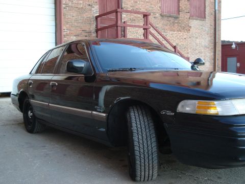 1992 Ford Crown Victoria - A black 1992 Ford Crown Victoria is parked in front of a brick building with a large garage door.