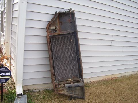 1965 Pontiac Catalina - A rusty car radiator assembly, likely removed from a vehicle, rests against a white vinyl-sided wall.