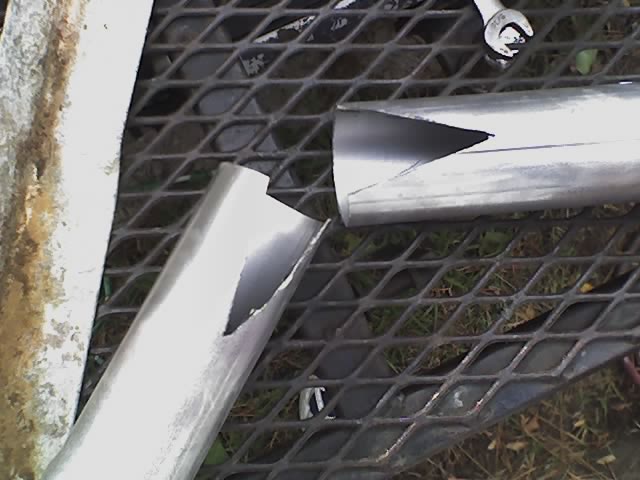 Two silver metal tubes with fishmouth-notched ends resting on expanded steel mesh grating over grass