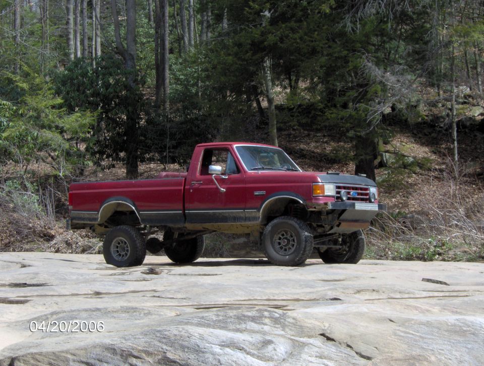Red 1990 Ford F-150 pickup truck parked on rocks in a wooded area