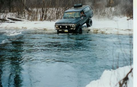 1978 Ford Bronco - A dark 1978 Ford Bronco navigates through a partially frozen river, kicking up spray.