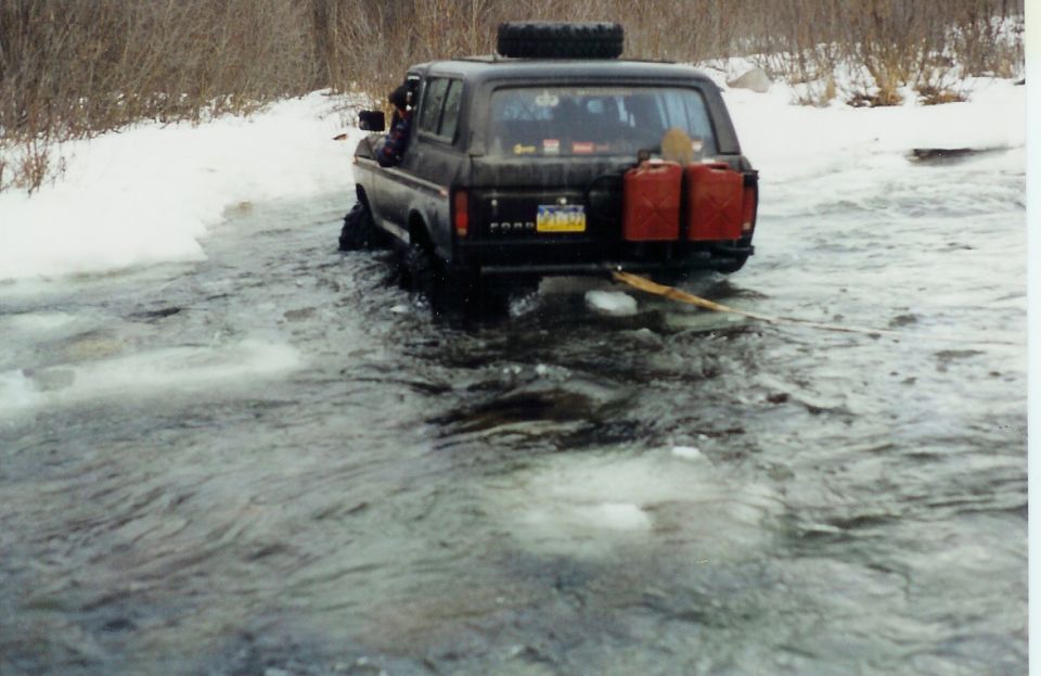 1978 Ford Bronco driving through icy water with two red jerry cans on the back and a spare tire on the roof