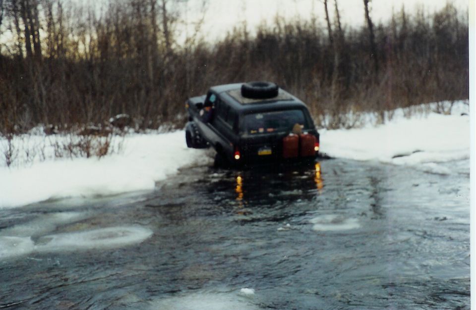 Black SUV with spare tire on roof and two gas cans on rear bumper driving through icy water with snow on banks