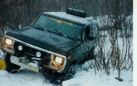 1978 Ford Bronco - A black 1978 Ford Bronco is stuck in a snowy, wooded area, with a winch attached and a spare tire on its roof.
