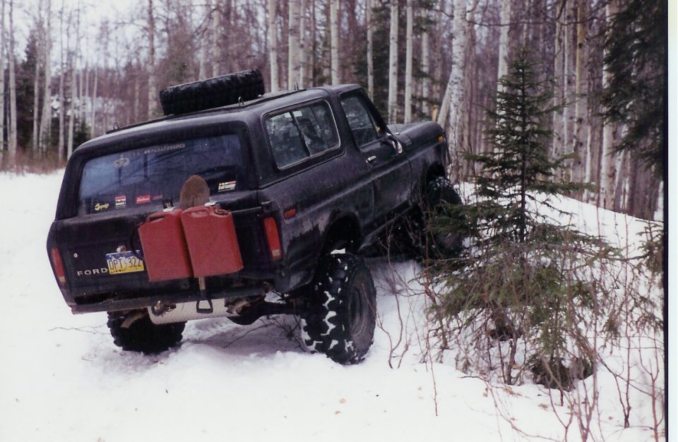 Black 1978 Ford Bronco with two red jerrycans and a shovel mounted on the rear, driving in snow