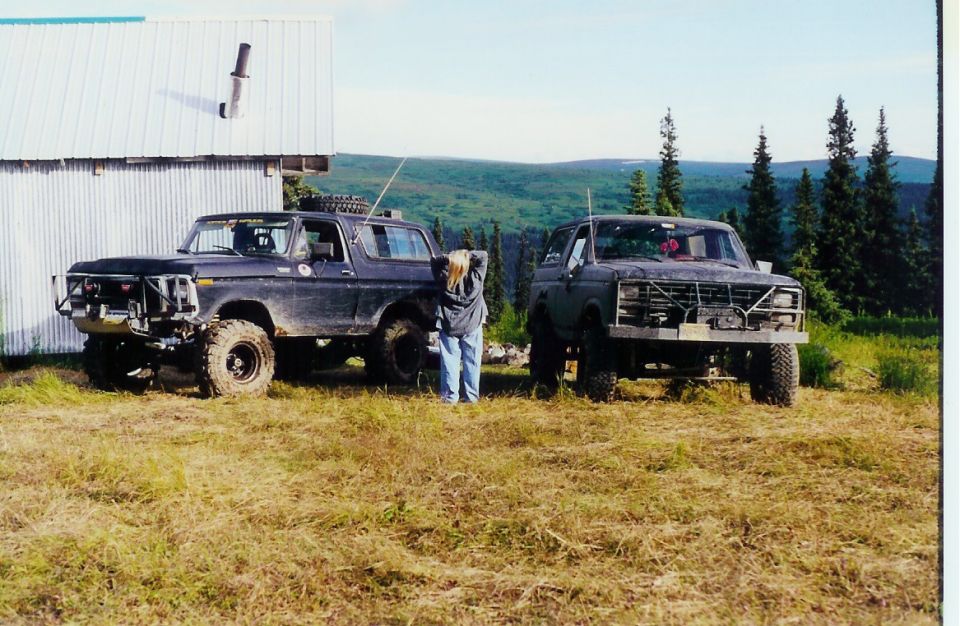 Two Ford Broncos parked in a grassy field near a building, with a person standing between them
