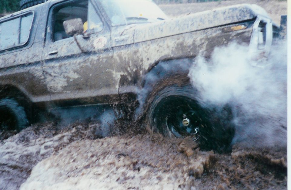 Mud-covered 1978 Ford Bronco spinning its front wheel in thick mud, kicking up spray