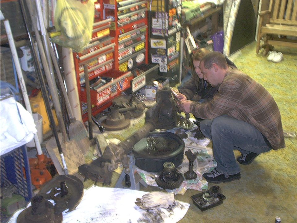 Two people are working on a disassembled vehicle differential in a garage, with tools and parts spread out