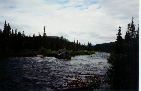 1978 Ford Bronco - An SUV navigates a rocky riverbed surrounded by dense forest under a cloudy sky.